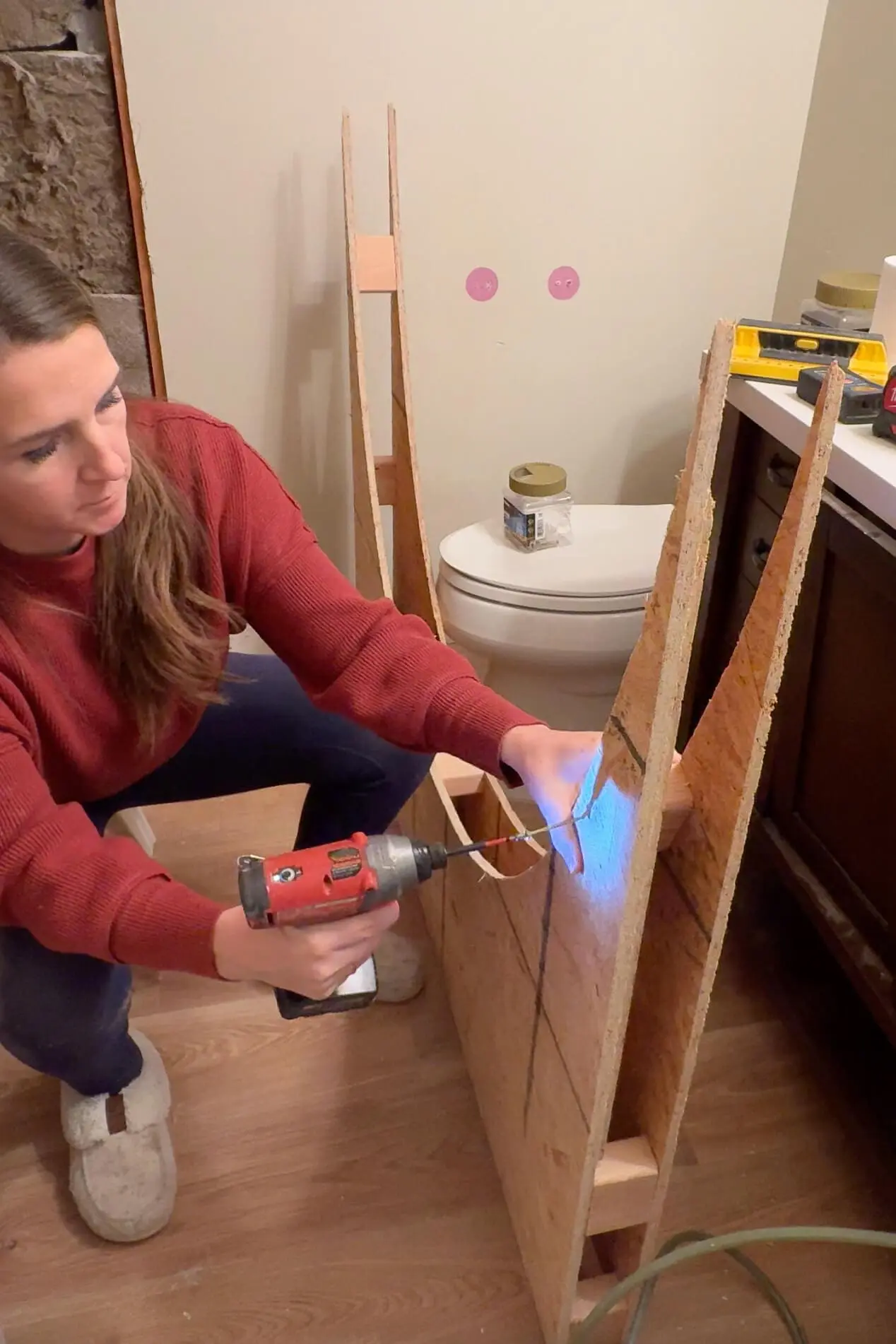A woman in a red sweater uses a power drill to secure pieces of an arched bathtub alcove. The structure, made from plywood and wood blocks, rests on a wooden floor in what appears to be an interior workspace.